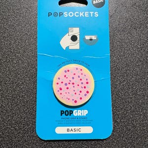 May include: A blue PopSockets package with a pink PopGrip featuring a pattern of colorful dots. The package includes instructions and the words "POPGRIP" and "BASIC". It is a phone grip and stand.