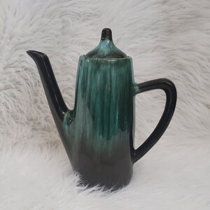 May include: A ceramic teapot with a long spout and handle. The teapot is glazed in shades of green and black, with a glossy finish. The lid has a small knob. The teapot is likely vintage or retro.