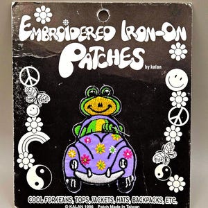 May include: Embroidered iron-on patch featuring a frog driving a purple car with flower accents. The patch is on a black background with peace signs, flowers, and butterflies. Text reads "COOL FOR JEANS, TOPS, JACKETS, HATS, BACKPACKS, ETC."