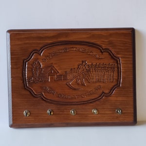 May include: A rectangular wooden key holder with a dark brown finish. The key holder features an engraved design with the words "GIVE US THIS DAY OUR DAILY BREAD" and a rural scene. It has five brass key hooks.
