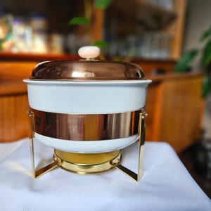 Peut inclure: Un chafing dish blanc et cuivré avec un couvercle et une base dorée. Le plat a une bande de cuivre au milieu et une petite poignée ronde sur le dessus. Le chafing dish est conçu pour servir des aliments.