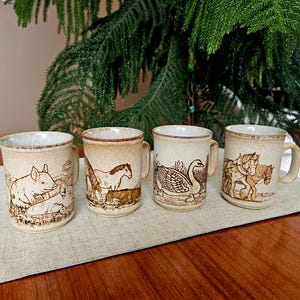 May include: Four beige ceramic mugs with brown animal illustrations. The mugs depict a pig, horses, a swan, and horses pulling a carriage. Each mug has a handle and a textured rim, ideal for a farmhouse or rustic kitchen.