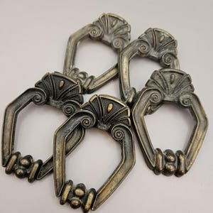 Peut inclure: Quatre poignées de tiroir antiques de couleur bronze. Chaque poignée présente un dessus décoratif en forme d'éventail avec des détails en volutes et une poignée incurvée. Les poignées sont disposées sur une surface blanche.