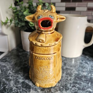 May include: A novelty ceramic cow-shaped container with a wide open mouth. The container is tan with a red mouth, blue eyes, and a bell around its neck. The word "FABULOUS" is printed on the front, along with a Las Vegas-themed design.