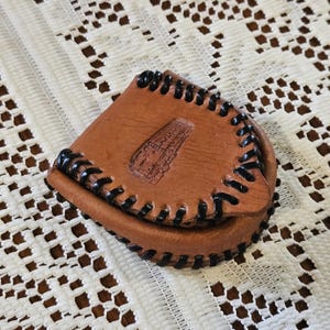 May include: A small, brown leather pouch with a flap closure. The pouch has black stitching around the edges and an embossed design on the front. The pouch is likely used for carrying small items.