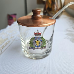 May include: A clear glass sugar bowl with a copper-coloured lid and wooden knob. The bowl features the Royal Canadian Mounted Police emblem. A small spoon rests inside the bowl.
