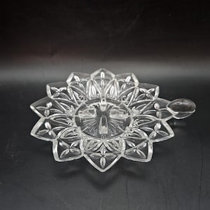 May include: Clear glass sun-shaped dish with a handle. The dish has a detailed, starburst-like design with a sun face in the centre. The handle is a small, rounded shape. The dish is on a dark background.