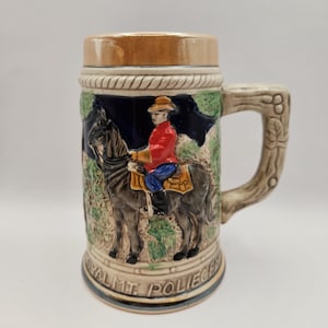 Peut inclure: Une chope à bière en céramique avec un relief détaillé d'une figure montée en veste rouge et chapeau marron. La chope a un dessus marron clair et une anse en forme de branche d'arbre. Les mots "ROYAL MT. POLICE" sont à la base.