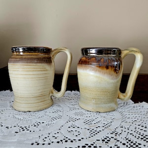 May include: Two handmade ceramic mugs with a unique, rustic design. The mugs feature a light beige base with brown and dark brown accents. Each mug has a large, curved handle and a glossy finish, perfect for tea or coffee.