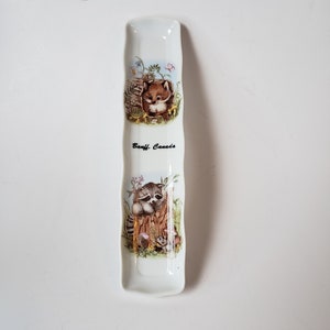 May include: White ceramic spoon rest with a wavy design. Features illustrations of a fox and a raccoon in a natural setting. The text "Banff, Canada" is printed on the rest.