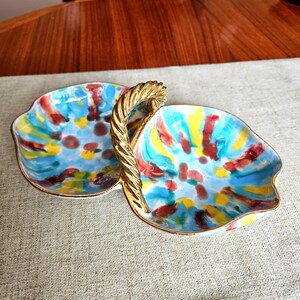 May include: A two-sectioned, hand-painted ceramic serving dish with a gold handle. The dish features a vibrant abstract design with blue, yellow, and red brushstrokes. The rim is trimmed with gold.