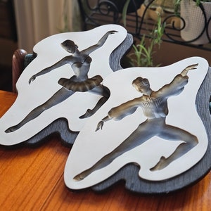 Peut inclure: Deux plaques murales décoratives représentant des danseurs de ballet en mouvement. Les figures sont dans des tons de gris et de marron, sur fond blanc. Les plaques ont un bord ondulé gris foncé, créant un effet de superposition. Le thème général est la danse et l'art.