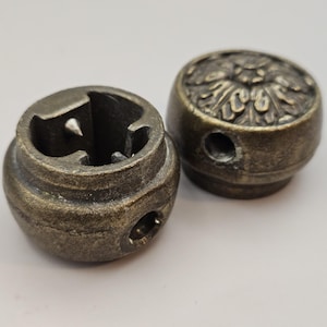 May include: Two antique bronze-coloured furniture knobs. One knob has a detailed floral design on the top, while the other has a hollowed-out interior with a small hole. Both knobs have a weathered appearance.