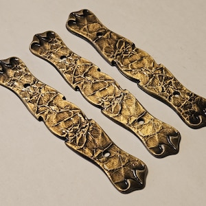 May include: Four antique-style, decorative metal plates with a textured, crumpled appearance. The plates are a metallic gold colour with a dark, aged finish. Each plate has a series of holes for mounting and decorative ends.