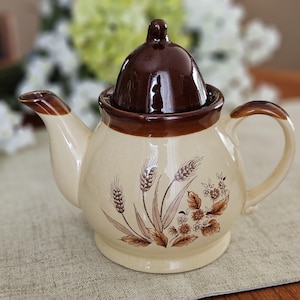Peut inclure: Théière en céramique crème avec couvercle et bordure marron. La théière présente un motif de blé et de fleurs dans des tons bruns. Elle possède une anse incurvée et un bec verseur.