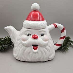 May include: A white ceramic teapot in the shape of Santa Claus, with a red hat, rosy cheeks, and a red nose. The handle is white with red stripes. The teapot has a whimsical design, ideal for festive decorations.