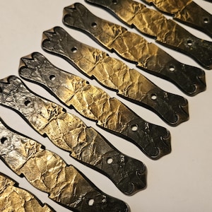 May include: A collection of decorative metal hardware. Each piece features a dark grey, ornate end and a gold-coloured, textured centre. The pieces are arranged diagonally, with a small hole near the centre of each.