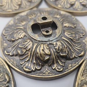 May include: A collection of antique brass-coloured metal decorative hardware. The circular pieces feature detailed floral and leaf patterns with a central mounting bracket. The aged finish adds to the vintage aesthetic. Suitable for furniture restoration.