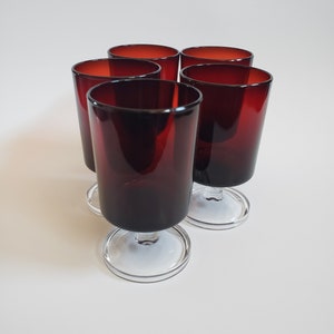 May include: Five ruby red stemmed glasses. The glasses have a cylindrical shape with a clear, circular base. The interior of the glasses is a deep red, transitioning to a darker shade towards the base. The glasses are arranged in a slightly offset circular pattern.