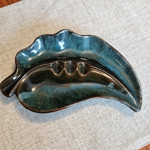 May include: A leaf-shaped ashtray with a glossy, two-tone finish. The ceramic ashtray features a dark teal and brown colour scheme with three cigarette rests. The ashtray has a unique, organic design.