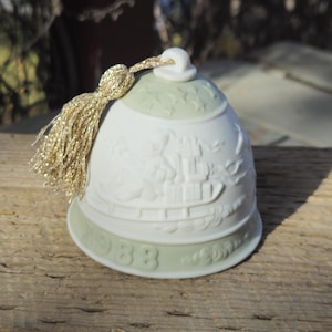 May include: A white ceramic bell with a gold tassel. The bell has a light green top and base, with a detailed relief design of a ship and figures. The year "1988" is inscribed on the base.