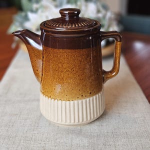 May include: A vintage ceramic coffee pot with a lid. The pot has a gradient of brown and tan colours, with a cream-coloured, vertically ribbed base. The handle and spout are brown. This retro coffee pot is a kitchenware item.