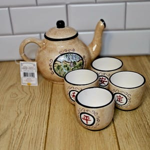 May include: A beige ceramic tea set featuring a teapot and four teacups. The teapot has a black handle and spout, and a black knob on the lid. The teacups have black rims and red Chinese characters. The teapot has a landscape painting.