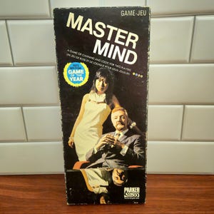 May include: A vintage "Master Mind" board game box. The box displays the game title in large white letters, with the text "A GAME OF CUNNING AND LOGIC FOR TWO PLAYERS". The box also features an image of a man and woman, and a "Game of the Year" sticker.
