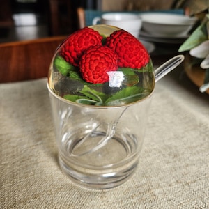 May include: A clear glass sugar bowl with a lid featuring three red raspberries and green leaves encased in clear resin. A clear spoon rests inside the bowl. The bowl sits on a beige linen surface.