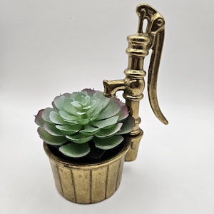 May include: A brass-effect water pump planter with a faux succulent. The plant has green leaves with red edges. The planter is a small, detailed replica of a vintage water pump, adding a touch of rustic charm.