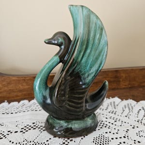 May include: A ceramic swan ornament with a glossy finish. The swan has a dark brown head and neck, blending into a teal and green body. The base is a matching green. This decorative piece is likely vintage.