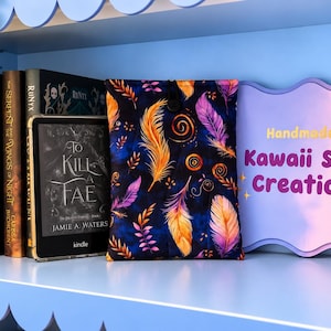 May include: A fabric e-reader sleeve featuring a dark blue background with a vibrant pattern of orange, yellow, and purple feathers and swirling lines, secured with a black button closure, positioned next to a Kindle and a sign reading Handmade by Kawaii Style Creations.