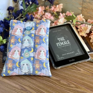 Peut inclure: Une housse de Kindle matelassée bleue avec un motif de cochons d'Inde portant des bonnets de nuit, des ours en peluche, des lunes et des étoiles. Une liseuse Kindle avec le titre du livre "THE FEMALE" est à côté de la housse. Des fleurs décoratives sont en arrière-plan.