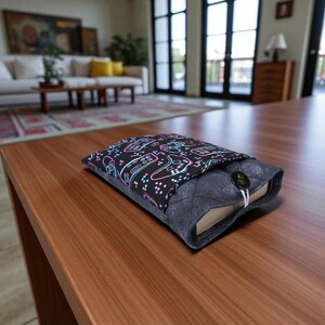 May include: A book sleeve with a black cover patterned with neon video game controllers. The quilted grey sleeve has a button closure. The book rests on a wooden table.
