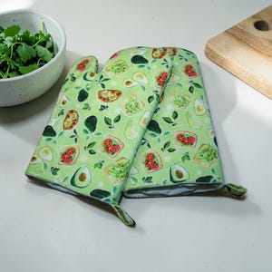 May include: A pair of green oven mitts with a pattern of avocado, toast, and herbs. A white bowl of fresh cilantro and a wooden cutting board are also in the image. The mitts are made of a soft, durable fabric.