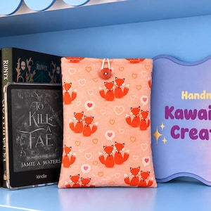 May include: A fabric e-reader sleeve featuring a repeating pattern of orange foxes and white hearts on a light peach background, secured with a small red button and white elastic loop, positioned next to a Kindle and a sign reading Handmade by Kawaii style Creations.