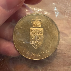 Op de afbeelding: Een gouden munt met een kroon- en schildontwerp. De munt heeft de tekst "CANADA" onderaan.