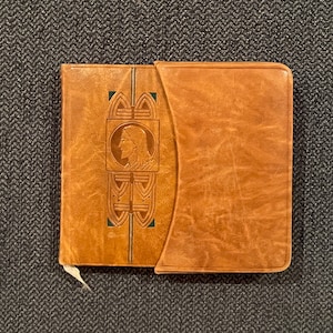 May include: A brown leather-bound book with a decorative design featuring a profile of a person with a crown of thorns. The design is in green and gold.