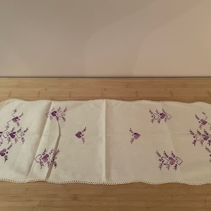 May include: A white table runner with a scalloped edge and purple embroidered floral designs.