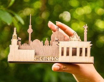 Könnte beinhalten: Ein Holzmodell der Berliner Skyline mit ikonischen Sehenswürdigkeiten wie dem Brandenburger Tor und dem Reichstagsgebäude. Das Modell wird in einer Hand vor einem verschwommenen grünen Hintergrund gehalten.