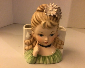 May include: A vintage ceramic planter shaped like a girl's head with blonde hair, a green dress, and a white flower in her hair. The planter has a rectangular opening in the back for planting.