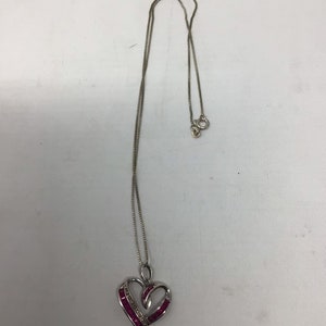 May include: A silver chain necklace with a heart-shaped pendant. The pendant is decorated with a row of pink gemstones and smaller clear gemstones.