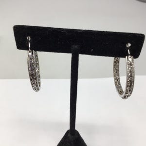May include: A pair of silver hoop earrings with a delicate, intricate design. The earrings are on a black display stand.
