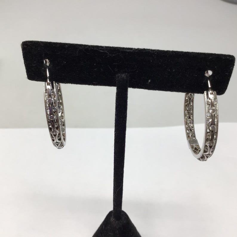 May include: A pair of silver hoop earrings with a delicate, intricate design. The earrings are on a black display stand.