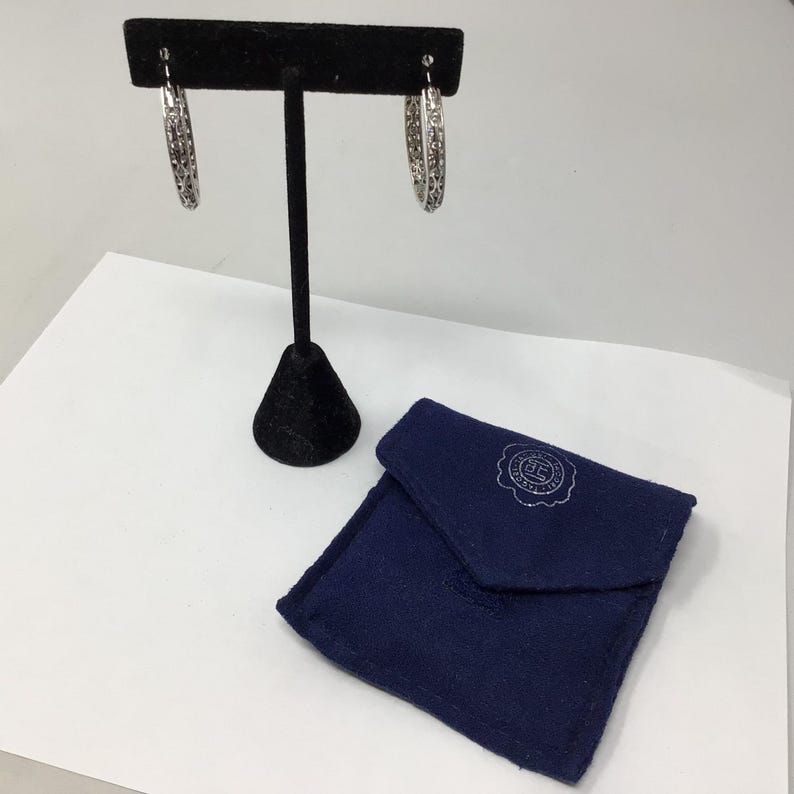 May include: A pair of silver hoop earrings with a delicate, intricate design. The earrings are displayed on a black stand and come with a blue velvet pouch.
