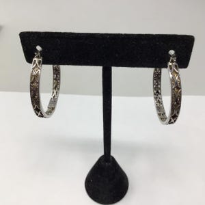May include: A pair of silver hoop earrings with a delicate, intricate design. The earrings are displayed on a black stand.