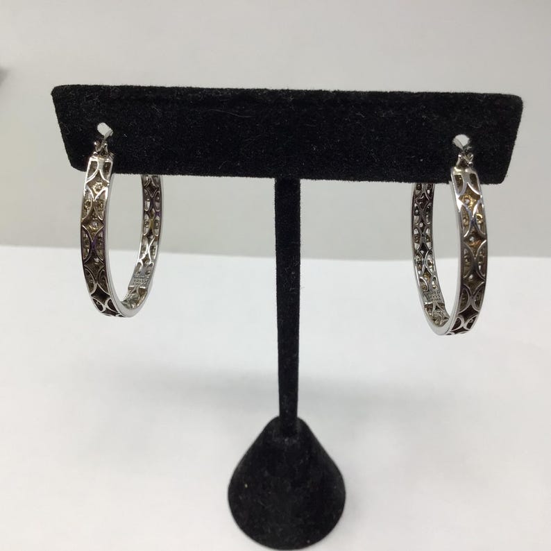 May include: A pair of silver hoop earrings with a delicate, intricate design. The earrings are displayed on a black stand.