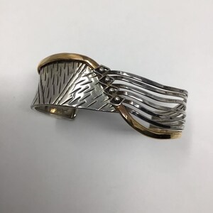 May include: A silver and gold cuff bracelet with a textured design and multiple bands. The bracelet is open and has a curved shape.