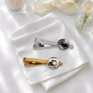 May include: Two tie clips, one silver-toned and one gold-toned, each featuring a circular photo insert. The silver clip is engraved with the text "Father of the Bride," while the gold clip is engraved with the text "Always your little girl."