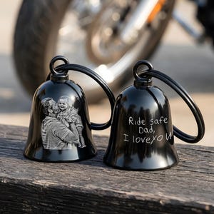 May include: Two black motorcycle bells with a loop for hanging. One bell features an engraved image of a man holding a child. The other bell has the text "Ride safe Dad, I love you".